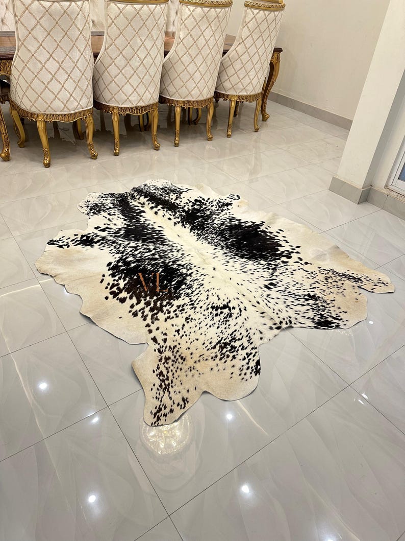 May include: A black and white cowhide rug with a natural, spotted pattern. The rug is laid out on a light grey tiled floor.