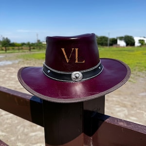 Puede incluir: Un sombrero vaquero de cuero burdeos con una banda negra y una concho plateada. El sombrero tiene las iniciales "VL" grabadas en la corona.
