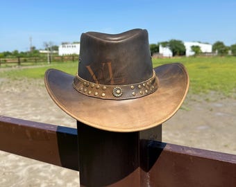May include: A brown leather cowboy hat with a distressed finish. The hat has a brown leather band with gold-toned studs and a metal buckle. The hat is sitting on a wooden fence post.