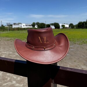 Könnte beinhalten: Ein brauner Cowboyhut aus Leder mit einem braunen Lederband und einer goldenen Metallschnalle. Der Hut hat die Initialen "VL" auf der Vorderseite geprägt.