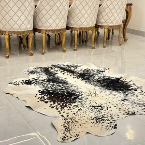 May include: A black and white cowhide rug with a natural, spotted pattern. The rug is laid out on a tiled floor in front of a set of gold-painted dining chairs.