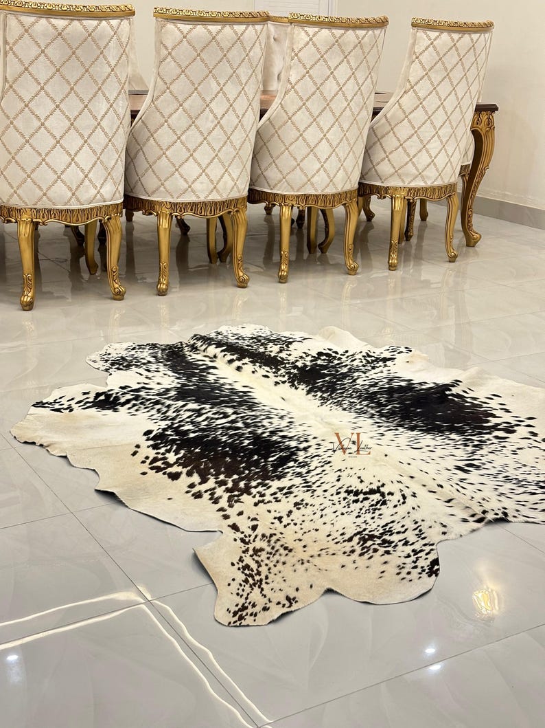 May include: A black and white cowhide rug with a natural, spotted pattern. The rug is laid out on a tiled floor in front of a set of gold-painted dining chairs.