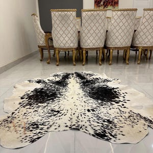 May include: A black and white cowhide rug with a spotted pattern. The rug is laid out on a tiled floor in front of a set of gold dining chairs.