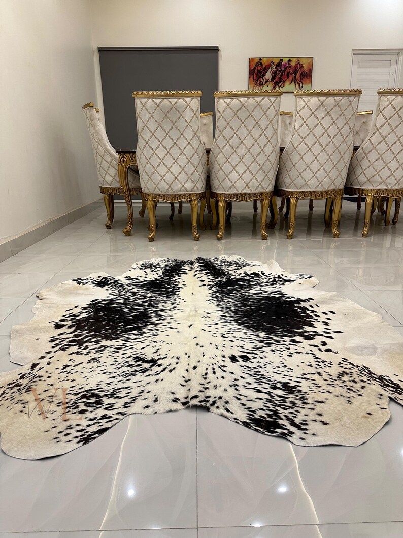 May include: A black and white cowhide rug with a spotted pattern. The rug is laid out on a tiled floor in front of a set of gold dining chairs.