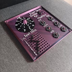 May include: A purple and black 3D printed tabletop game tracker with dials for hit points, death saves, modifiers, and ability scores. The tracker also includes a grid for spell slots.