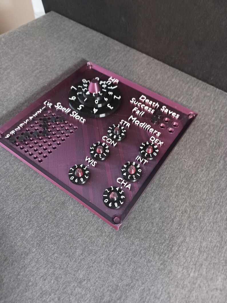 May include: A purple acrylic tabletop game board with a black dial for hit points, a grid for spell slots, and dials for ability scores, including strength, dexterity, constitution, wisdom, intelligence, and charisma. The board also includes a section for death saves with success and fail indicators.