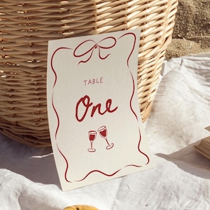 May include: A picnic setting featuring a wicker basket, a card reading "Table One", a book, and a tangerine. The card has a red border and text, with two wine glasses. The basket contains dried flowers.