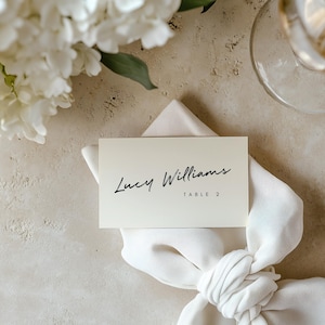 May include: A wedding table setting with a white napkin tied in a knot, a place card that reads "Lucy Williams, Table 2", a glass of white wine, and white flowers. The background is a neutral beige.