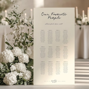 May include: A cream-colored seating chart with the words "Our Favorite People" in elegant script. Below, the text "please find your seat!" is printed. The chart lists names under table numbers. White flowers and candles are in the background.