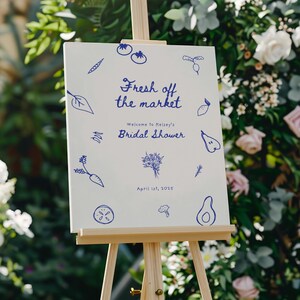 May include: A white sign with blue illustrations of vegetables and fruits, announcing a bridal shower for Kelsey on April 1st, 2025. The sign reads "Fresh off the market" and "Welcome to Kelsey's Bridal Shower".