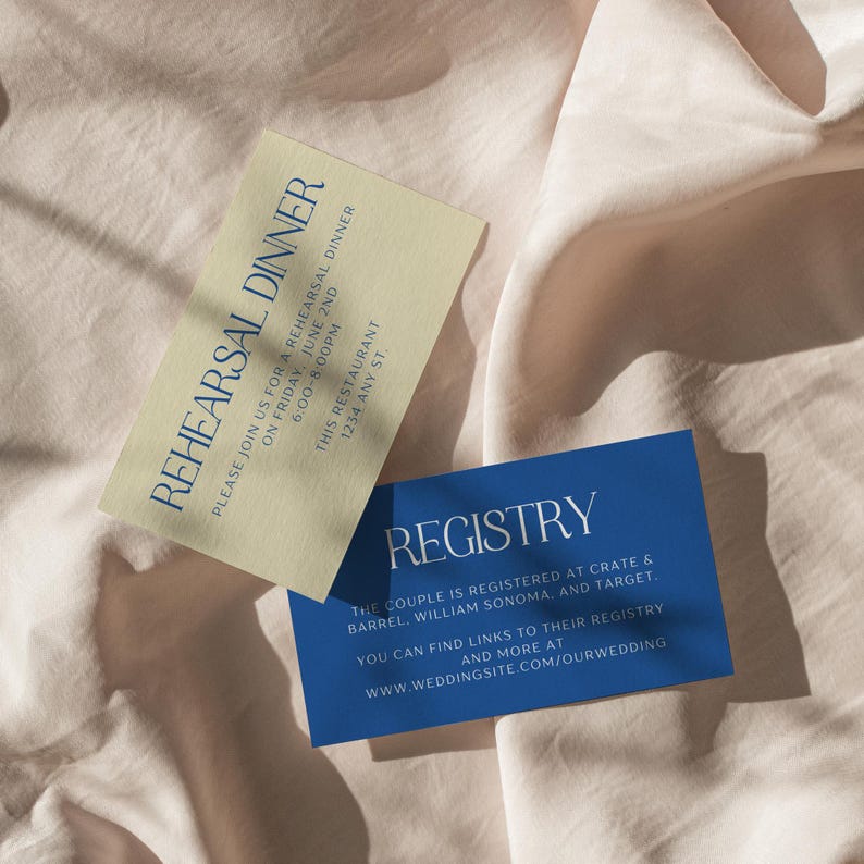 May include: Two cards with blue and gold text on a beige fabric background. The first card is gold with blue text that reads "Please join us for a rehearsal dinner on Friday, June 2nd 6:00-8:00PM This Restaurant 1234 Any St.". The second card is blue with white text that reads "Registry The couple is registered at Crate & Barrel, William Sonoma, and Target. You can find links to their registry and more at www.weddingsite.com/ourwedding".