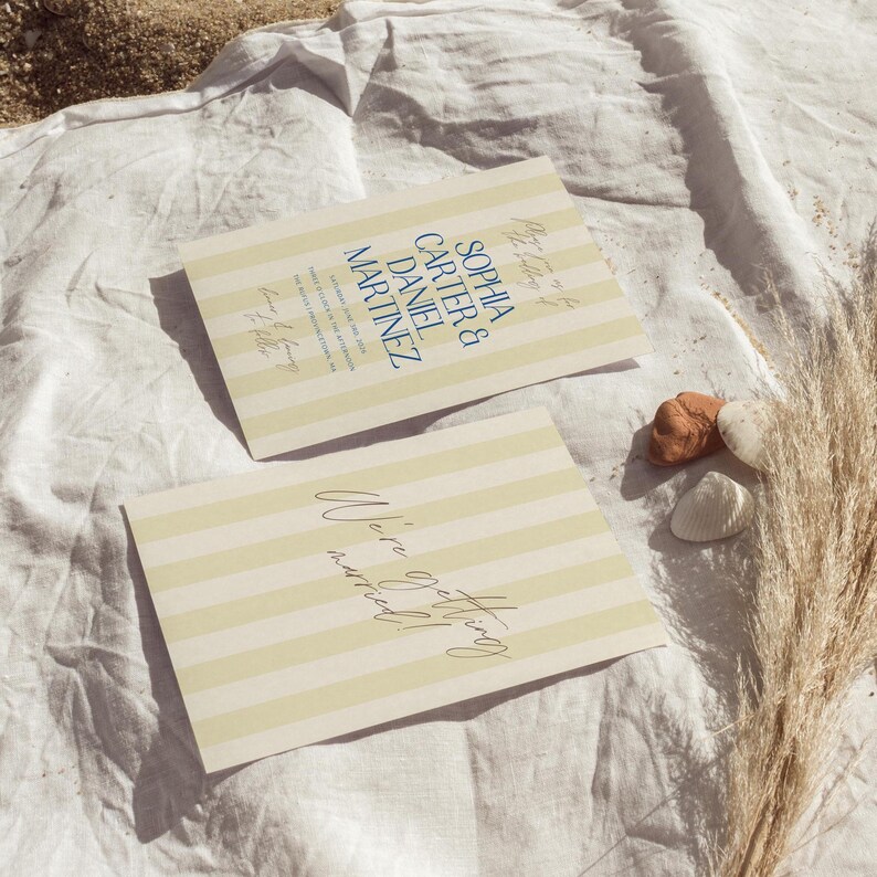 May include: A yellow and white striped card with the text "We're getting married!" in blue cursive writing. The card is lying on a white fabric surface.