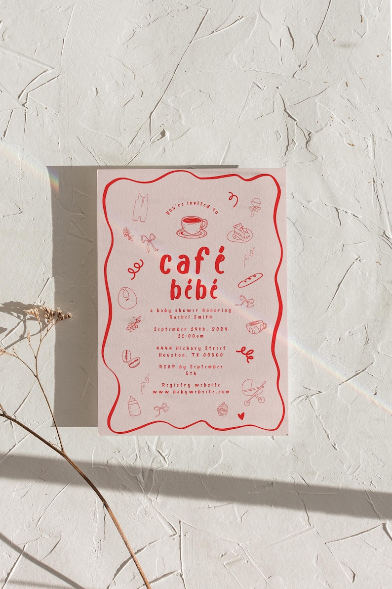 A 5x7 invitation card lying on a textured white surface. The invitation is for a baby shower with a French coffee shop theme called "café bébé". The fonts are whimsical and handwritten, and there are silly hand drawn illustrations all over.