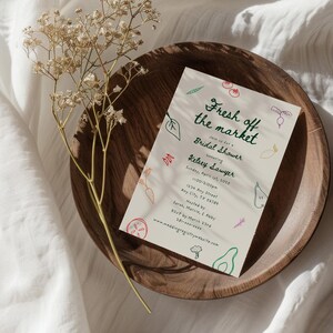 May include: A white invitation card with green text that reads "Fresh off the market" and "Bridal Shower honoring Kelsey Sawyer". The card is decorated with colorful illustrations of vegetables and fruits.