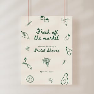 May include: A white sign with green illustrations of vegetables and fruits. The text reads "Fresh off the market" and "Welcome to Kelsey's Bridal Shower" with the date "April 1st, 2025".