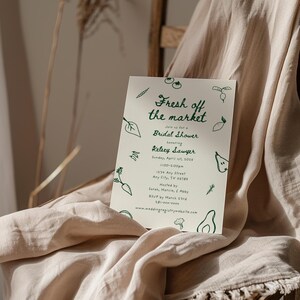 May include: A white card with green text and illustrations of vegetables and fruits. The text reads "Fresh off the market" and "Join us for a Bridal Shower honoring Kelsey Sawyer". The card also includes the date, time, address, and RSVP information.