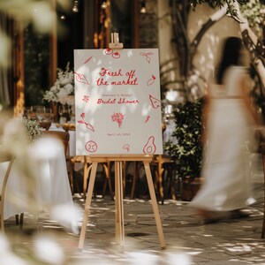 May include: A white sign with red text and illustrations of produce, announcing a bridal shower. The sign reads "Fresh off the market" and "Welcome to Hailey's Bridal Shower" with the date "April 1st, 2023". The sign is on a wooden easel.
