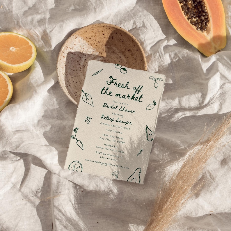A 5x7 bridal shower invitation card lying on a white linen cloth outside in the sun. There's oranges and papaya next to it. The invite's design features whimsical hand drawn illustrations of fruit and veggies, with the title 'fresh off the market'.