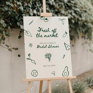 A 18x24 welcome sign for a bridal shower with the theme "Fresh off the market". The sign is resting on a wood easel. The design features hand drawn doodles of fruit and veggies, and the fonts look handwritten. The aesthetic is modern and whimsical.