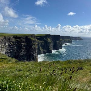 May include: A panoramic view of the Cliffs of Moher in Ireland. The cliffs are tall and rugged, with green grass growing on top. The ocean is blue and choppy, with white waves crashing against the rocks. The sky is blue and cloudy.
