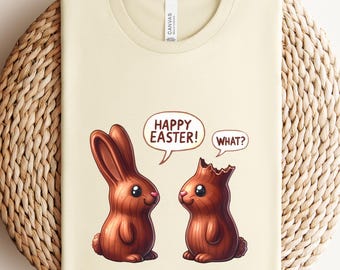 May include: A cream-colored t-shirt featuring a cartoon illustration of two chocolate bunnies. One bunny says "Happy Easter!" while the other responds with "What?" The shirt is folded on a woven, circular mat.