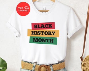 May include: White t-shirt with the text "BLACK HISTORY MONTH" in black letters on three colored stripes: red, yellow, and green. The shirt is being modeled on a person wearing denim shorts and a brown belt.