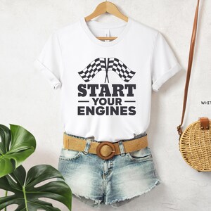 May include: White t-shirt with a black and white checkered flag graphic and the text "START YOUR ENGINES". The shirt is paired with denim shorts and a tan belt. A woven purse hangs on the right.