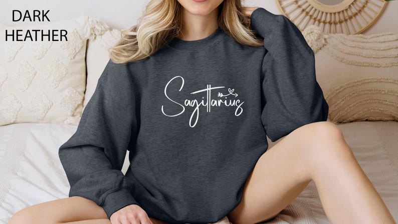 K&ouml;nnte beinhalten: Ein dunkel-heathergrauer Pullover mit dem Schriftzug "Sagittarius" in wei&szlig;er Schrift. Der Schriftzug ist in einer kursivschriftlichen Schriftart gehalten und enth&auml;lt ein kleines Pfeil- und Herzdesign.