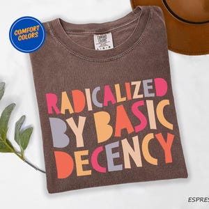 May include: Brown t-shirt with the text "Radicalized by Basic Decency" in colorful letters. The text is in a retro style font.