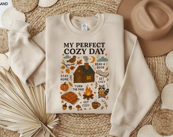 May include: A sand-colored sweatshirt with the text "MY PERFECT COZY DAY" and illustrations of a cabin, bird, pumpkin, and raccoon. The design includes the phrases "Stay Home", "Turn the Page", "Read a Book", and "Get Cozy".