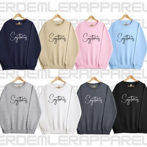 K&ouml;nnte beinhalten: Ein Set aus zehn Crewneck-Sweatshirts in verschiedenen Farben, jedes mit dem Text "Sagittarius" in einer handschriftlichen Schriftart. Die Sweatshirts sind in Marineblau, Beige, Rosa, Hellblau, Grau, Wei&szlig;, Dunkelgrau und Schwarz erh&auml;ltlich.