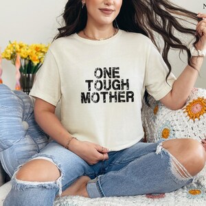May include: A white t-shirt with black text that reads "ONE TOUGH MOTHER". The shirt is being worn by a person sitting on a white crocheted blanket.