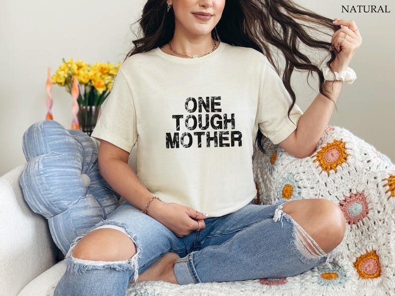 May include: A white t-shirt with black text that reads "ONE TOUGH MOTHER". The shirt is being worn by a person sitting on a white crocheted blanket.