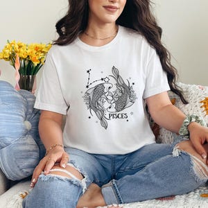 May include: White t-shirt featuring a black and gray Pisces zodiac design. The graphic includes two fish, floral elements, and the word "PISCES". The shirt is a casual style.