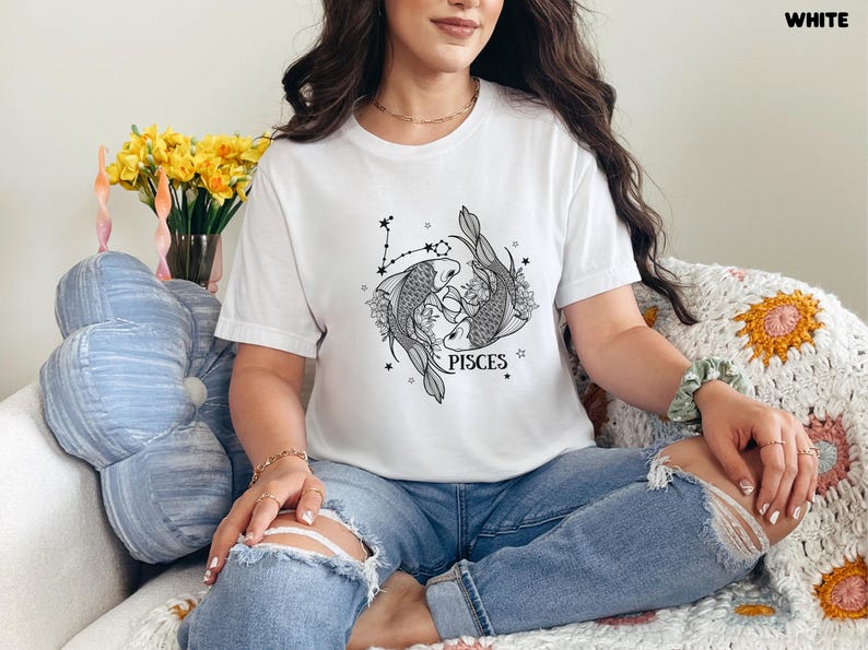 May include: White t-shirt featuring a black and gray Pisces zodiac design. The graphic includes two fish, floral elements, and the word "PISCES". The shirt is a casual style.