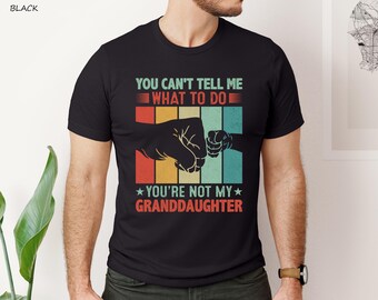 May include: Black t-shirt with a retro design. The graphic features two fists bumping, with text that reads "YOU CAN'T TELL ME WHAT TO DO YOU'RE NOT MY GRANDDAUGHTER" in red, teal, yellow, and orange. The shirt is a crew neck.