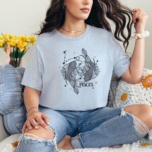May include: Light blue t-shirt featuring a Pisces design. The graphic shows two fish, floral elements, and stars, with the word "PISCES" below. The shirt is worn with blue jeans.