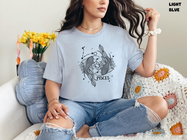 May include: Light blue t-shirt featuring a Pisces design. The graphic shows two fish, floral elements, and stars, with the word "PISCES" below. The shirt is worn with blue jeans.