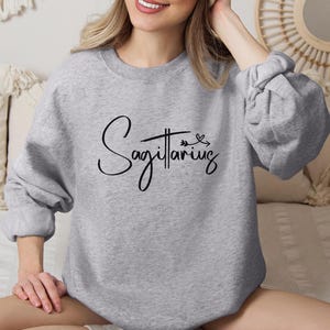 K&ouml;nnte beinhalten: Ein grauer Pullover mit dem Wort "Sch&uuml;tze" in schwarzer Kursivschrift. Der Pullover hat einen Rundhalsausschnitt und lange &Auml;rmel.