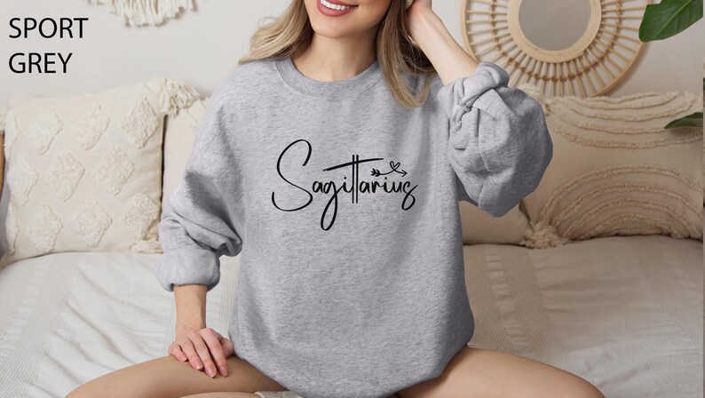 K&ouml;nnte beinhalten: Ein grauer Pullover mit dem Wort "Sch&uuml;tze" in schwarzer Kursivschrift. Der Pullover hat einen Rundhalsausschnitt und lange &Auml;rmel.