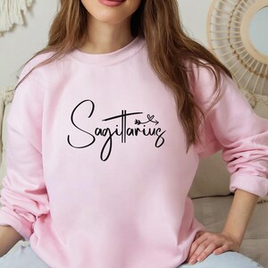 K&ouml;nnte beinhalten: Ein weicher, rosa Pullover mit dem Wort "Sch&uuml;tze" in schwarzer Kursivschrift.