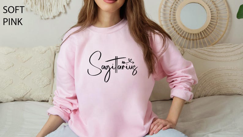 K&ouml;nnte beinhalten: Ein weicher, rosa Pullover mit dem Wort "Sch&uuml;tze" in schwarzer Kursivschrift.