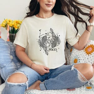 May include: Cream-colored t-shirt featuring a black and white Pisces graphic. The design includes two fish, constellations, and the word "PISCES". The shirt is worn with blue jeans.