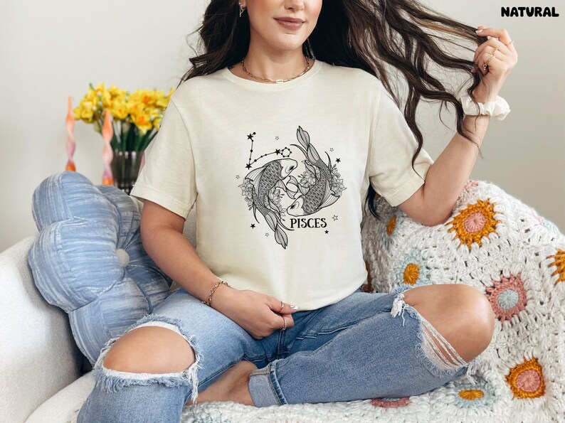 May include: Cream-colored t-shirt featuring a black and white Pisces graphic. The design includes two fish, constellations, and the word "PISCES". The shirt is worn with blue jeans.