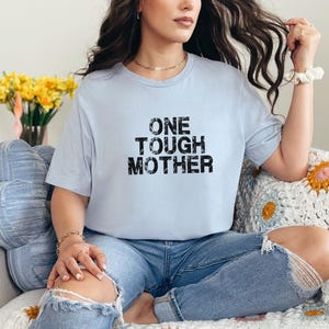 May include: A light blue t-shirt with the text "ONE TOUGH MOTHER" printed in black. The shirt is being worn by a person sitting on a couch.