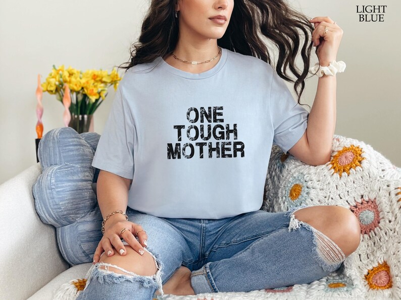 May include: A light blue t-shirt with the text "ONE TOUGH MOTHER" printed in black. The shirt is being worn by a person sitting on a couch.