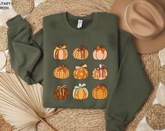 May include: Military green sweatshirt featuring a pattern of nine pumpkins with various designs and bows. The pumpkins are in shades of orange, brown, and peach. A tan hat is visible in the background. The sweatshirt is a fall-themed item.