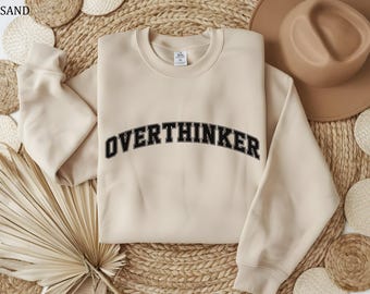 May include: A sand-colored sweatshirt with the word "OVERTHINKER" in black, arched lettering. The sweatshirt is made of a soft material and has a crew neck and long sleeves. The image is taken from a top-down perspective, with a hat and decorative items in the background.