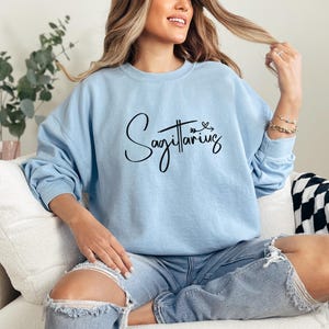K&ouml;nnte beinhalten: Ein hellblauer Pullover mit dem Schriftzug "Sagittarius" in schwarzer Schrift mit einem kleinen Herz und einem Pfeil &uuml;ber dem Wort.