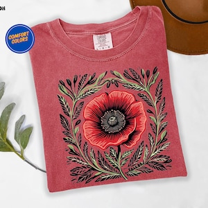 May include: Crimson red t-shirt featuring a large red poppy flower design with black and green leaves. The shirt has a crew neck and short sleeves. The words "Comfort Colors" are printed on a blue circle on the upper left corner.